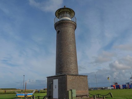 A tall brick lighthouse in a meadow with a few boats and a cloudy sky in the background, juist,