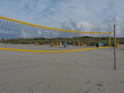A yellow volleyball net on the beach with several beach chairs and dunes under a cloudy sky, juist,