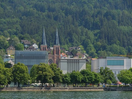 Two striking church towers and modern buildings in a green, wooded landscape on the lakeshore,