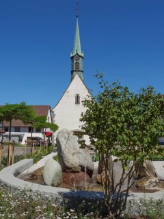 Church tower and fountain in a village surrounded by green trees and flowering plants on a sunny