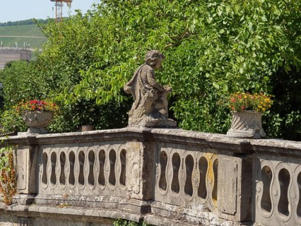 Stone statue on an old balcony railing, surrounded by green trees and plants, a touch of history