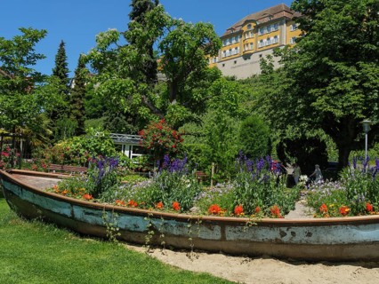 An old boat serving as a flower box in a green garden with a large yellow building in the