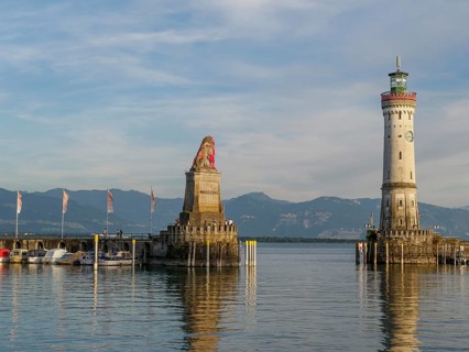 View of a harbour with a lion sculpture and a lighthouse against a mountain backdrop in the morning