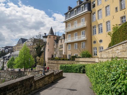 Curve of an urban street with yellow buildings and overgrown pavement, cloudy sky, Luxembourg City,