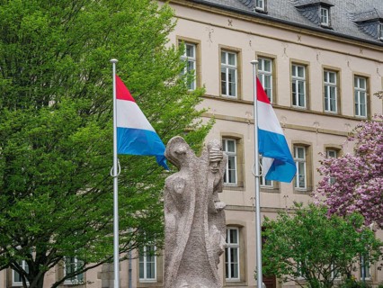 Monument with two Luxembourg flags in front of a historic building with blossoming trees,