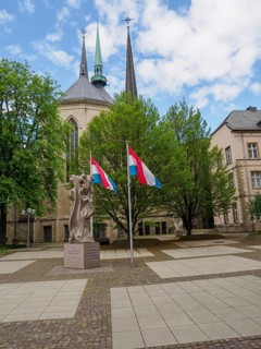 Modern square with a church, flags and a monument, surrounded by green trees, Luxembourg City,