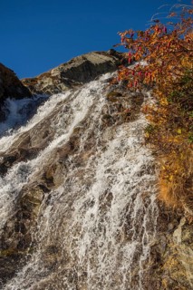 Water flows down a rocky slope, accompanied by colourful autumn leaves under a bright blue sky,