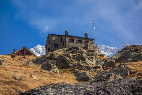 A mountain hut stands on a rocky slope with a distant snow-capped mountain range in the background,