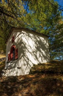A white chapel stands in the forest under sunlight, surrounded by trees and shade, Saas Fee,