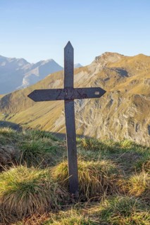 A rusty, iron cross on a mountain peak with an impressive mountain landscape and clear sky in the
