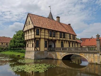 A half-timbered house with red roof tiles standing over a river with water lilies on a stone bridge