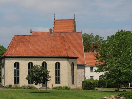 A small chapel with red roof tiles and large windows, surrounded by meadows and a tree, under a