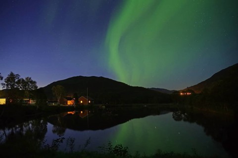 House and hill under the night sky with green northern lights and a reflection in the lake,