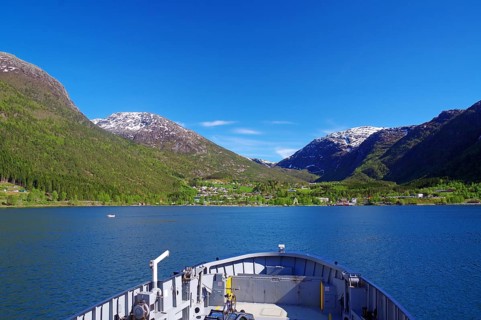 View from a ferry onto a Norwegian fjord surrounded by snow-capped mountains and blue sky,