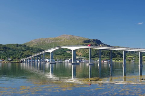 A curved bridge spans calm waters with reflective mountains and green landscapes under a clear sky,