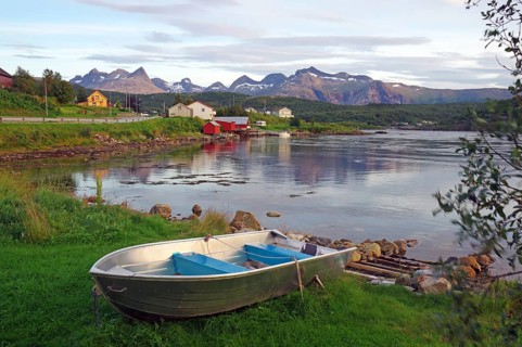 An abandoned boat lies on a grassy shore with mountains and houses in the background, Saltstraumen,