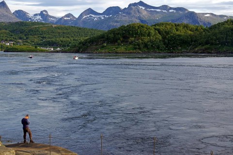 A man stands on the bank of a tidal stream, surrounded by green hills and mountains in the
