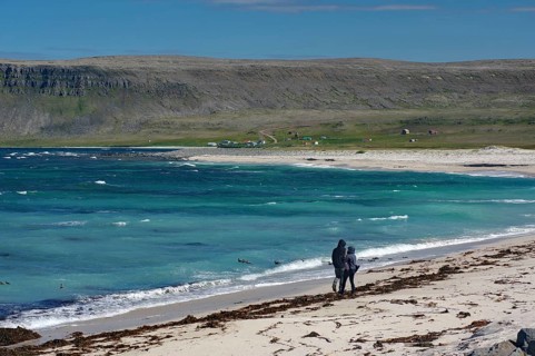 Two people walking along a beach with clear blue water and rocks in the background, Latrabjarg,