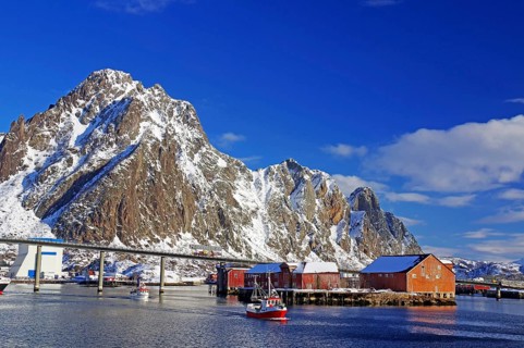 Harbour with bridge, surrounded by snowy mountains, boats in the water and sheds on the shore,