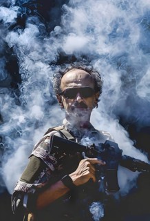 A mercenary in uniform and sunglasses stands in the powder smoke with his assault rifle, AI