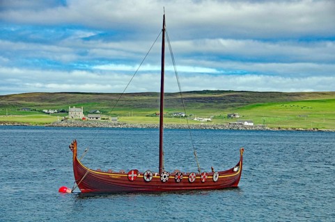 A traditional Viking boat with shields on the calm water, overlooking a green coastline and hills