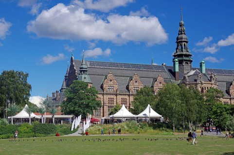 Historic building with striking architecture next to a green space and people under a clear sky,