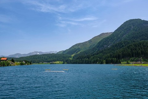 View of a lake with blue water, surrounded by green hills and forests under a clear sky with few