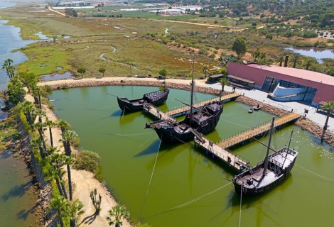 Historically modelled sailing ships in a calm water basin, surrounded by landscape under a clear