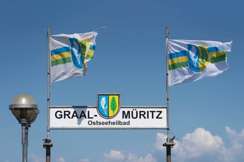 Two flags and a sign with the inscription Graal-Müritz Ostseeheilbad under a clear blue sky with
