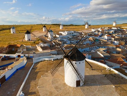 Many windmills in a rural setting with a village in the background under a cloudy sky. White houses