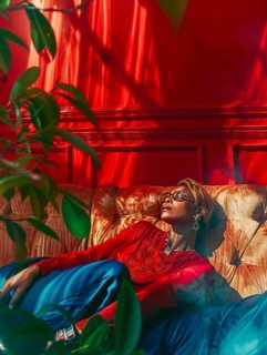 Stylish woman in a red and blue outfit lounging on a couch amidst plants and dramatic sunlight, AI