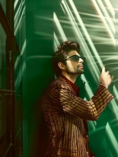 Stylish man in sunglasses and a striped jacket leaning against a green wall, lit by sunlight and