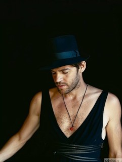 Man in a black hat and attire, wearing a necklace with red charm, appearing deep in thought under