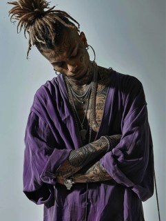 Man with dreadlocks and tattoos wearing a purple shirt, arms crossed and looking down in a moody
