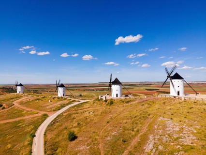 Several white windmills stand along a dirt road under a clear blue sky, aerial view, Alcazar de San