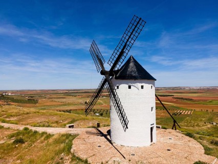 A white windmill with a black roof in front of a wide field landscape and blue sky, aerial view,