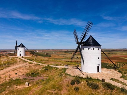 Two white windmills with black roofs in a vast landscape of fields under a sunny sky, aerial view,