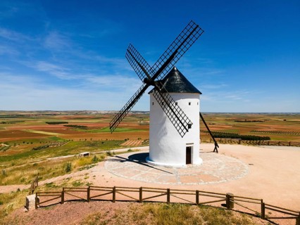 A white windmill with a black roof in the middle of fields under a clear blue sky, aerial view,