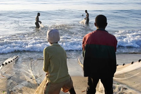 Boys are pulling a fishing net out of the water. Two fishermen are helping them. At Toamasina