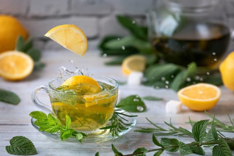 Lemon slices fall splashing into a glass of tea, surrounded by mint, sage and lemon halves