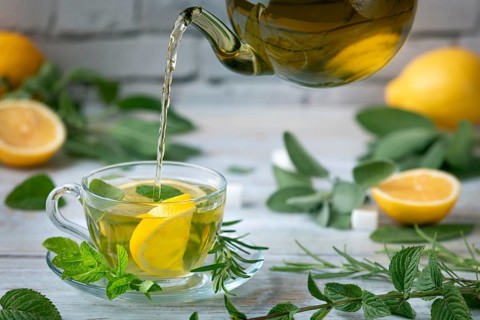 Tea is poured from a glass jug into a glass cup, surrounded by lemons and herbs