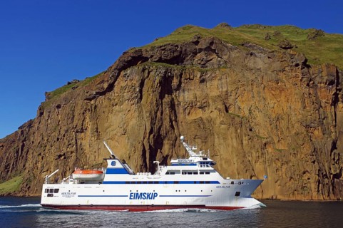 A passenger ship sails in front of a steep cliff in the blue sea, ferry to the Westman Islands,
