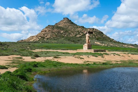 A statue stands in front of a grassy hill and a pond under a cloudy sky, Monument, Pastor
