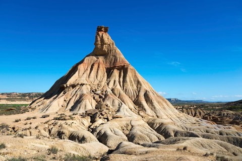 An imposing rock formation in the middle of a desert-like area under a light blue sky,