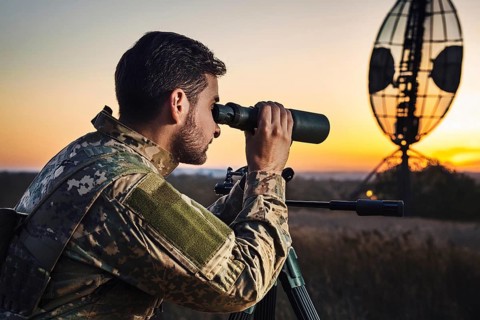 Man in military uniform using binoculars at sunset, with a radar in the background, AI generated,
