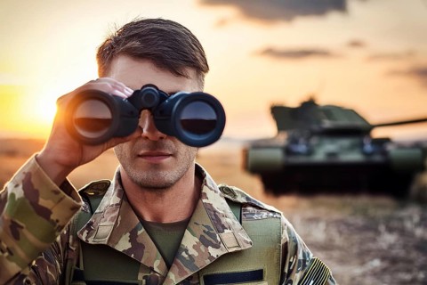 Man in camouflage holding binoculars, with a tank in the background and a dramatic sunset, AI