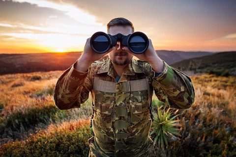 Man in camouflage clothing looks with binoculars over a green landscape at sunset, AI generated, AI
