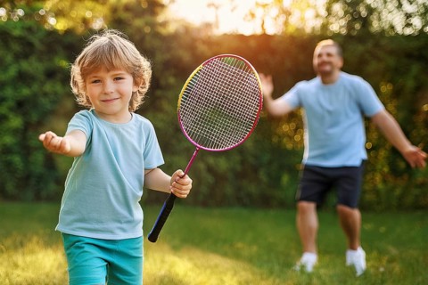 Smiling child in the garden playing with a badminton racket, in the background an adult person, ai