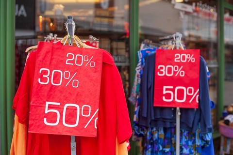 Red sign discount on a shopping bag in front of a clothing rack with t-shirts and shop window,