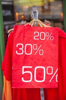 Red sign discount on a shopping bag in front of a clothing rack with t-shirts and shop window,